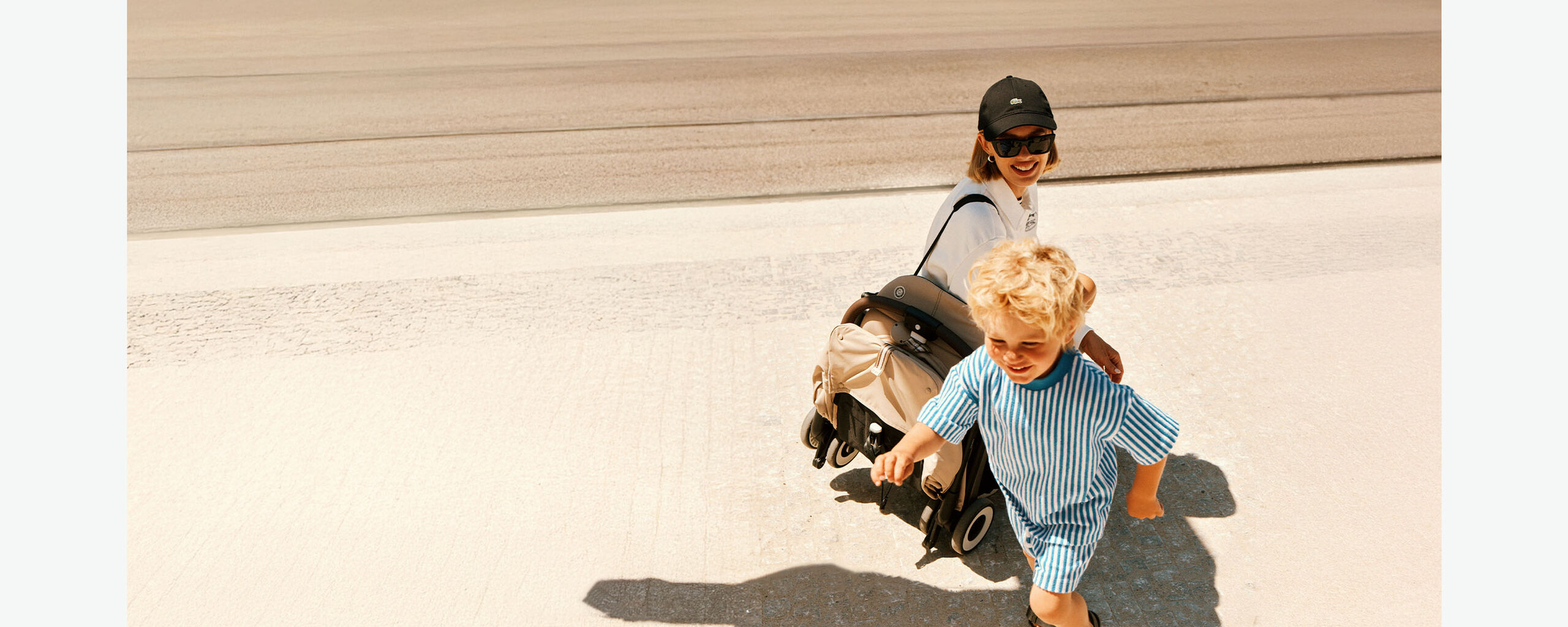 Eine Frau mit Sonnenbrille und Cap sieht ihrem Sohn dabei zu, wie er spielt. Sie hat einen zusammengeklappten CYBEX Buggy &uuml;ber ihre Schulter geschlungen.