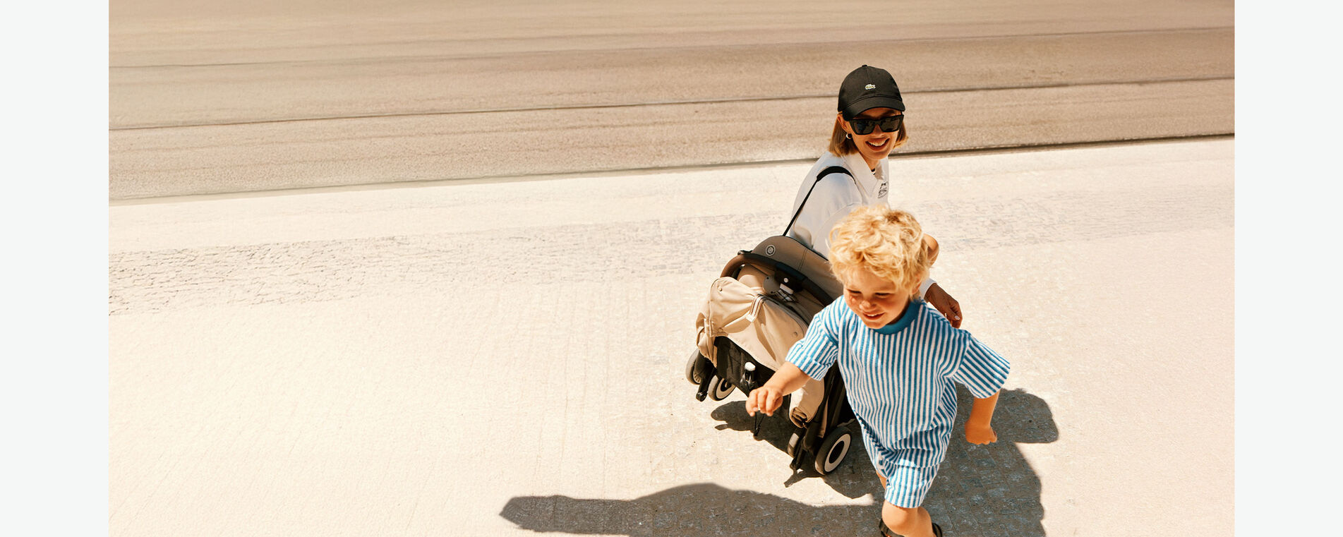 Eine Frau mit Sonnenbrille und Cap sieht ihrem Sohn dabei zu, wie er spielt. Sie hat einen zusammengeklappten CYBEX Buggy über ihre Schulter geschlungen.