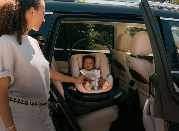 A woman standing at an open car door, holding the hand of her baby that's sitting in an Aton G2 Swivel car seat.
