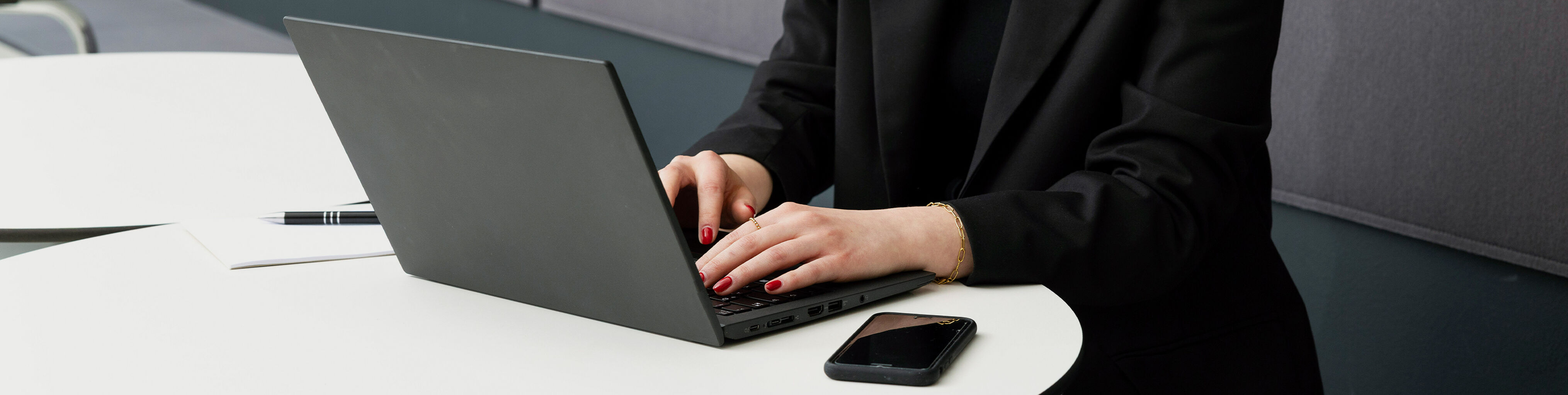 A person in a black blazer is typing on a laptop while a smartphone is resting on the table.
