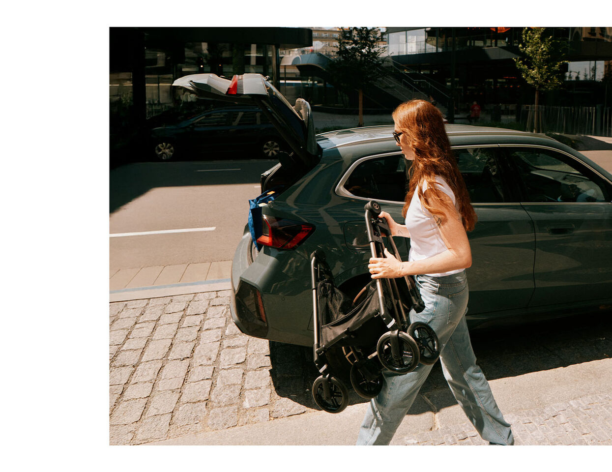 Frau mit brünetten, offenen Haaren geht auf ein Auto zu. Sie hält einen zusammengeklappten Kinderwagen in der Hand und ist dabei, ihn in den offenen Kofferraum zu laden. Die Szene spielt im Freien bei Sonnenschein. Sie trägt eine Jeans.