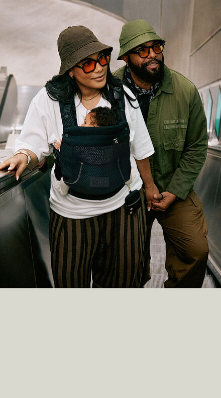 Woman carrying a baby in the black CYBEX Laya Carrier while riding an escalator alongside her partner.
