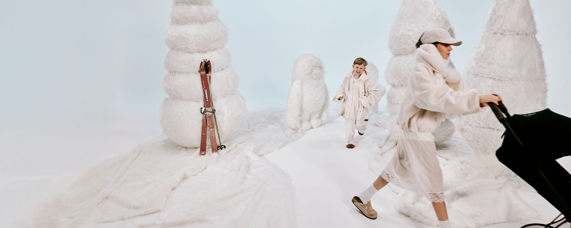 Deux enfants jouent au milieu d’un décor de neige improvisé, avec des skis appuyés sur la gauche. Un garçon court après une fille qui pousse une poussette CYBEX Priam.