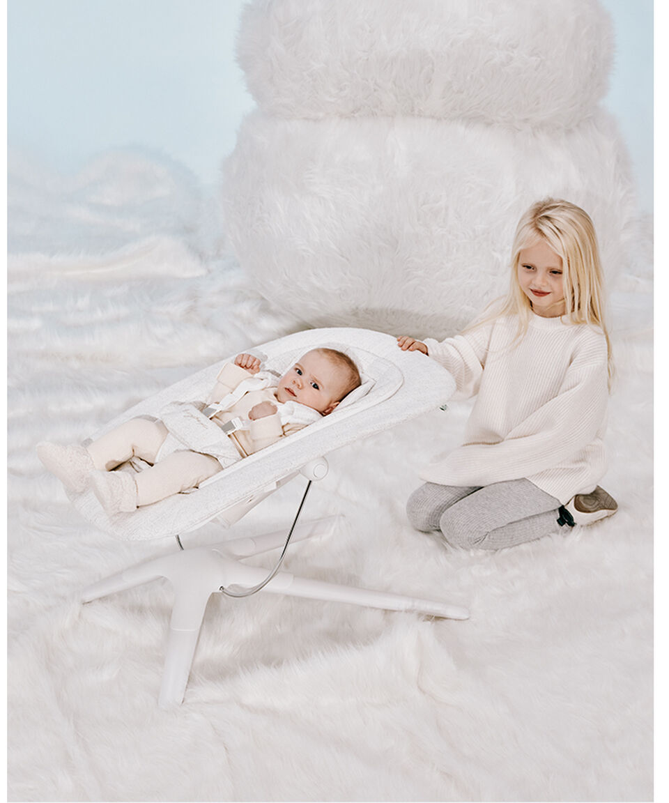 Une petite fille est assise derrière un transat dans lequel un bébé se repose, le tenant comme si elle l’aidait à le bercer.