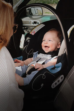 A child sitting inside an infant car seat, facing the door of the car, grinning at their mother.