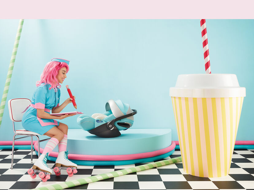 A woman with pink hair and roller skates sits on a chair, looking at a by Jeremy Scott Wings car seat in a retro-inspired setting with a giant striped cup in the foreground.