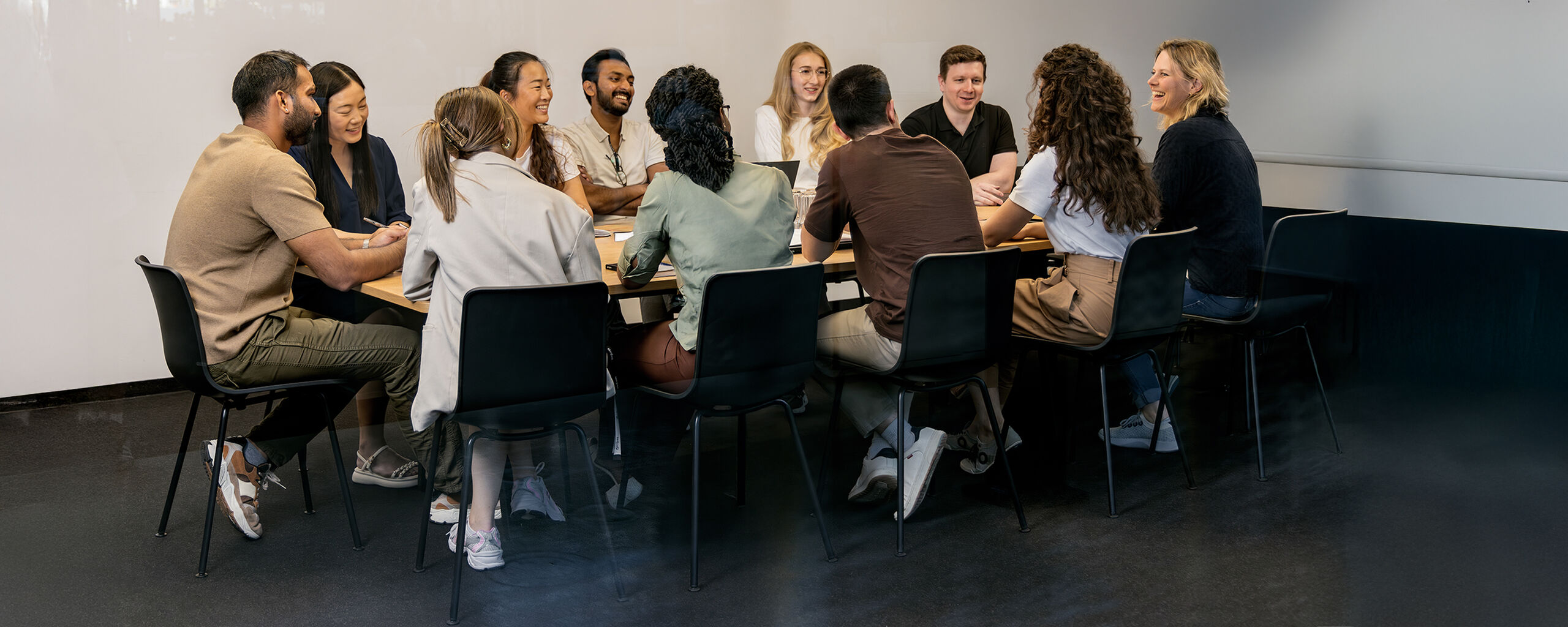 A group of eleven people is sitting around a table in a modern meeting room, engaging in lively discussion.