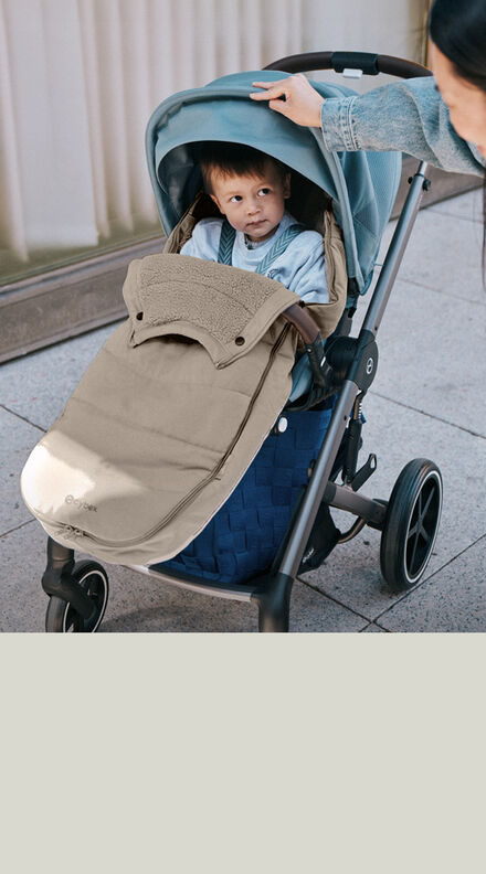 A child sitting in a CYBEX buggy and a Gold Footmuff keeping him warm. There's a hand at the sun canopy of the buggy.