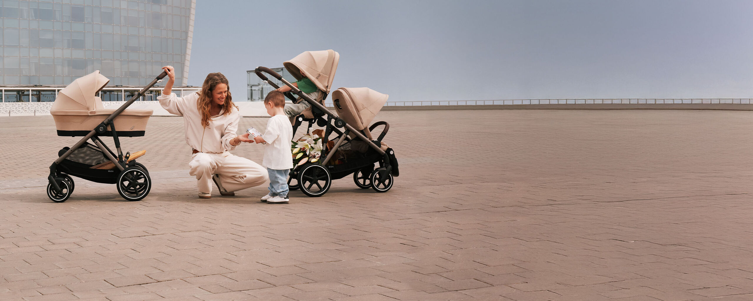 A mother is kneeling on the ground next to her child, grinning at him. She has two Gazelle S strollers next to her, one in single mode, one in sibling mode.