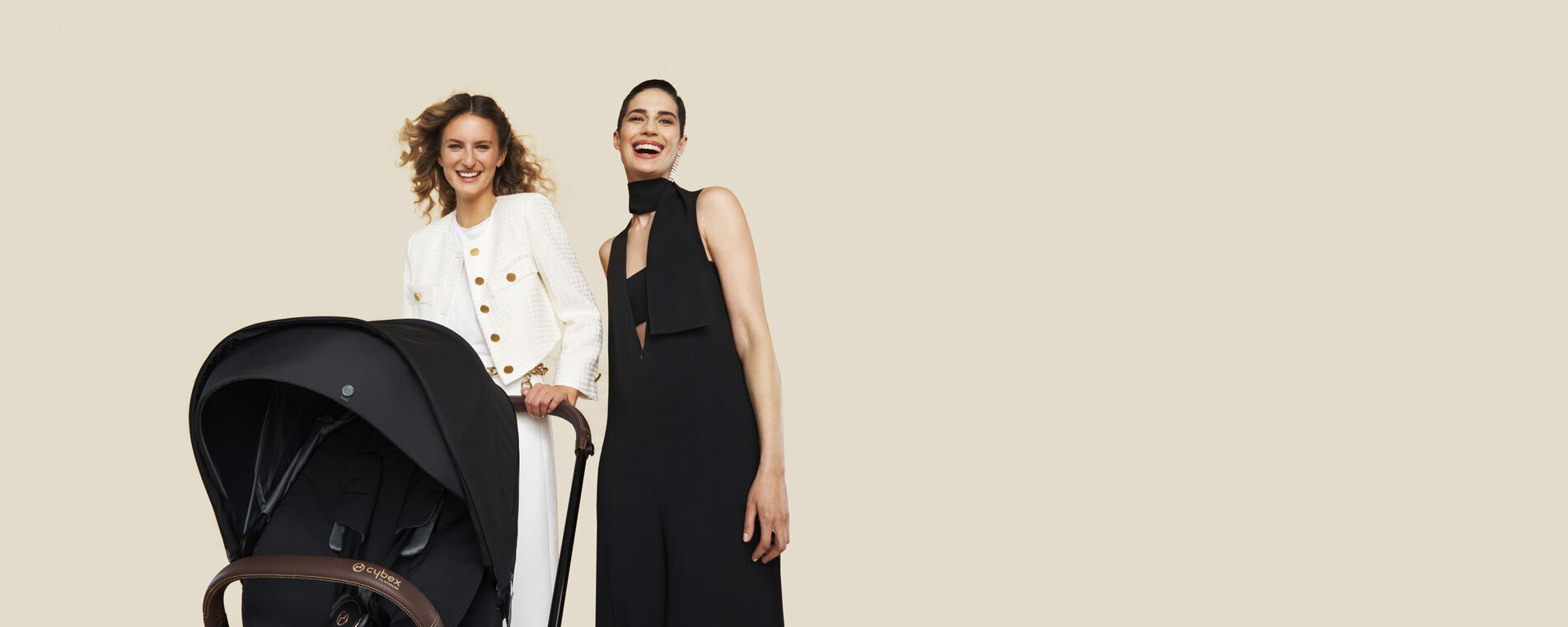 Zwei l&auml;chelnde Frauen, eine in einem wei&szlig;en Blazer und die andere in einem schwarzen Kleid, stehen neben einem schwarzen Kinderwagen von Cybex.