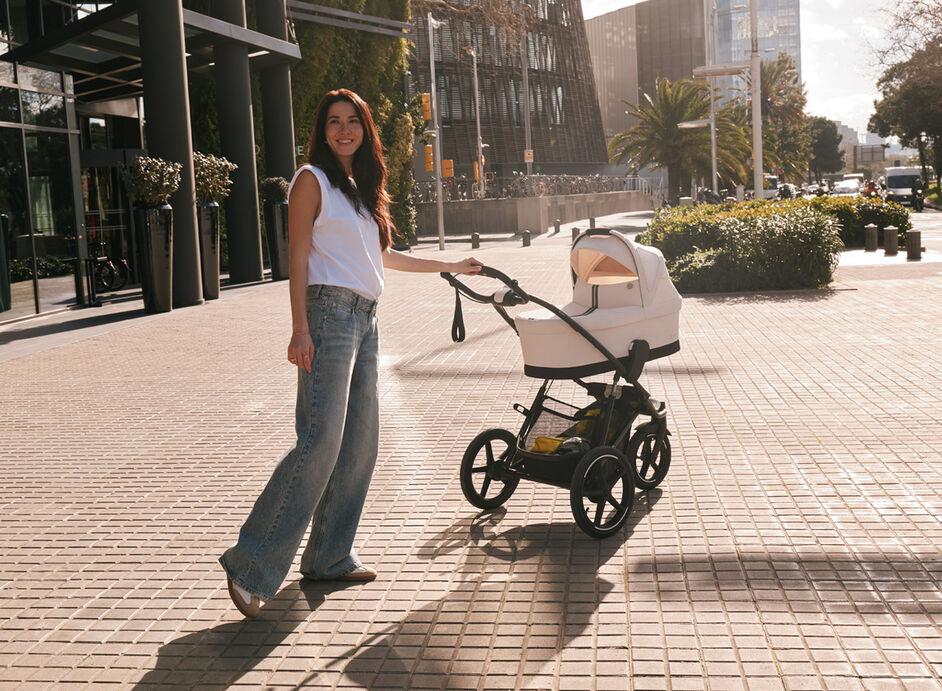A woman smiling into the camera with one hand on her Avi Spin stroller with a Cot attached.