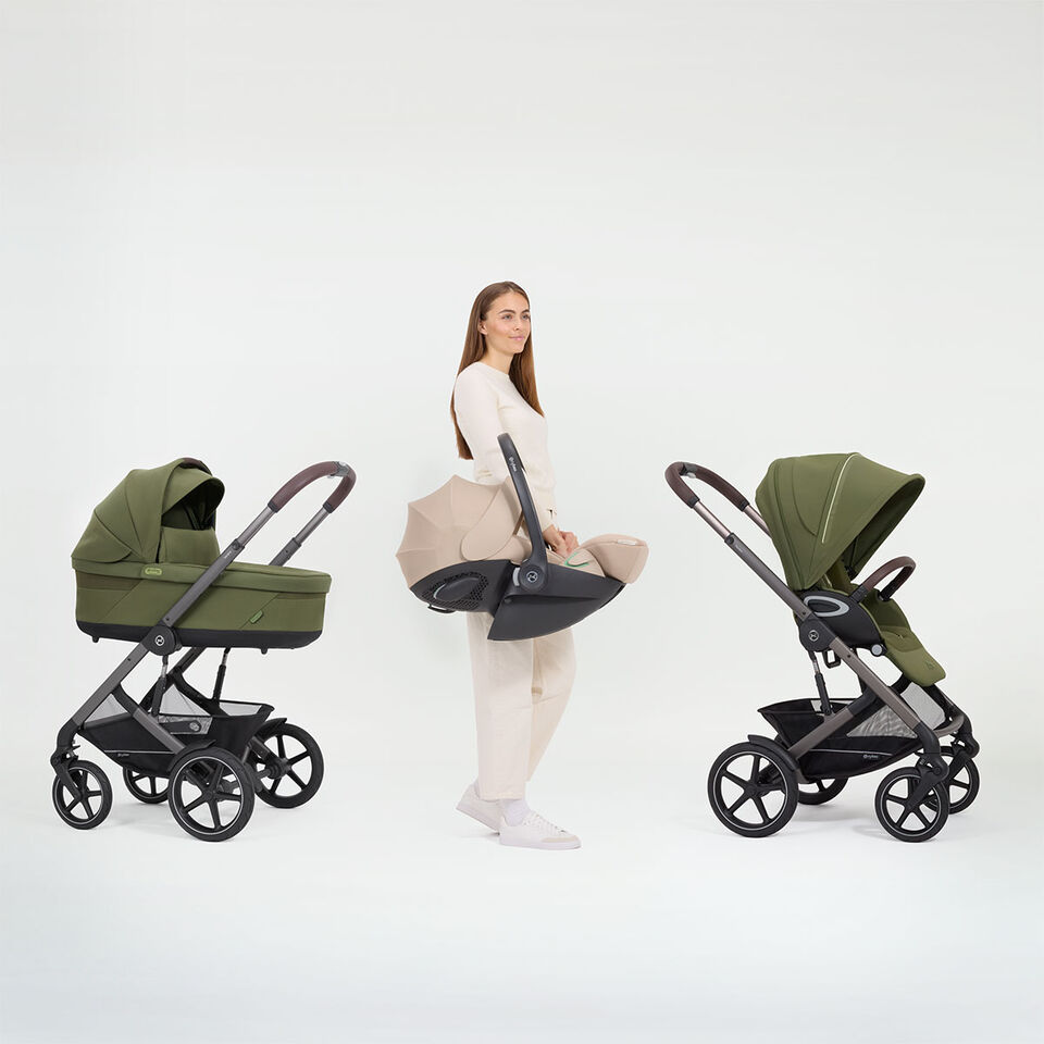 A woman standing between two Talos S Lux strollers, holding an infant car seat, showcasing the travel system ability the stroller has.