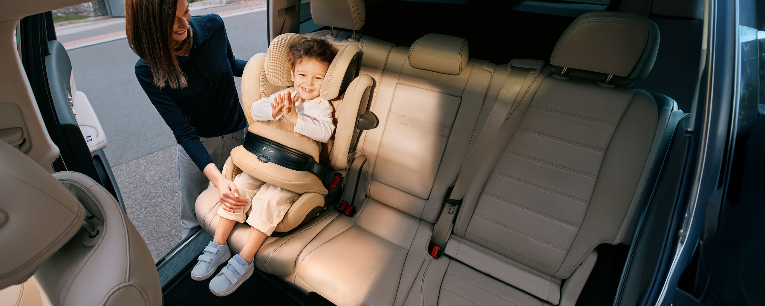 A child in the backseat of a car in a Pallas G3 car seat, clapping happily. The mother has one hand on their knee and is smiling at them.