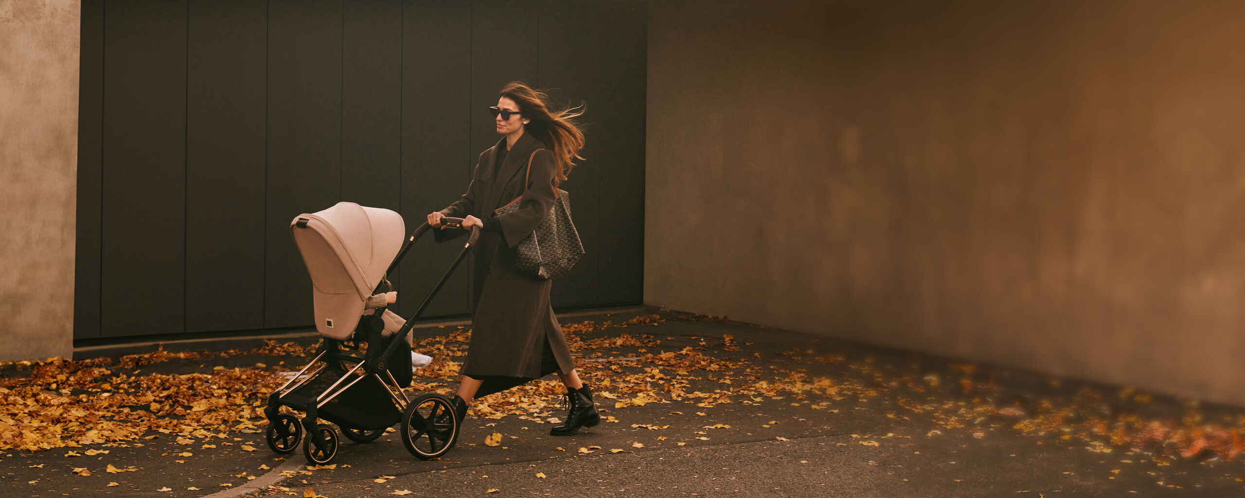 Woman pushing a pram on an autumn pavement, surrounded by fallen leaves.