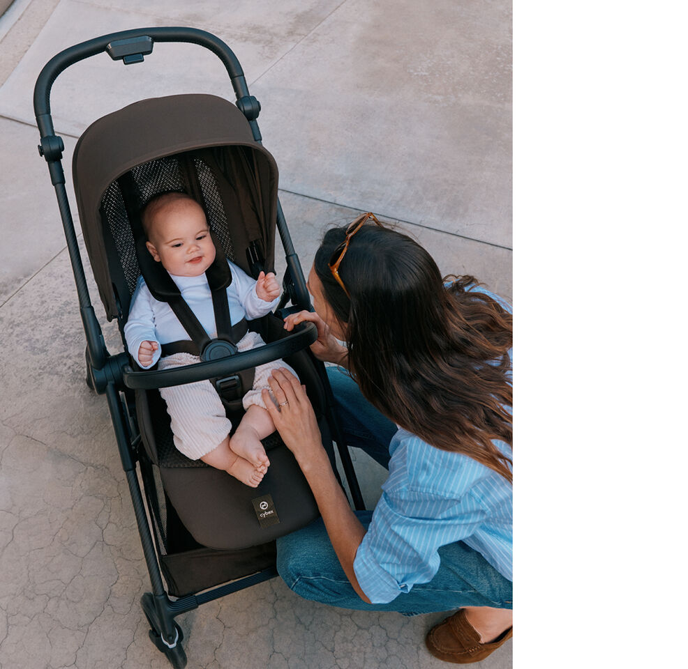 A baby sits in a Melio Carbon, the lightweight stroller, while a hand fastens the safety belt.