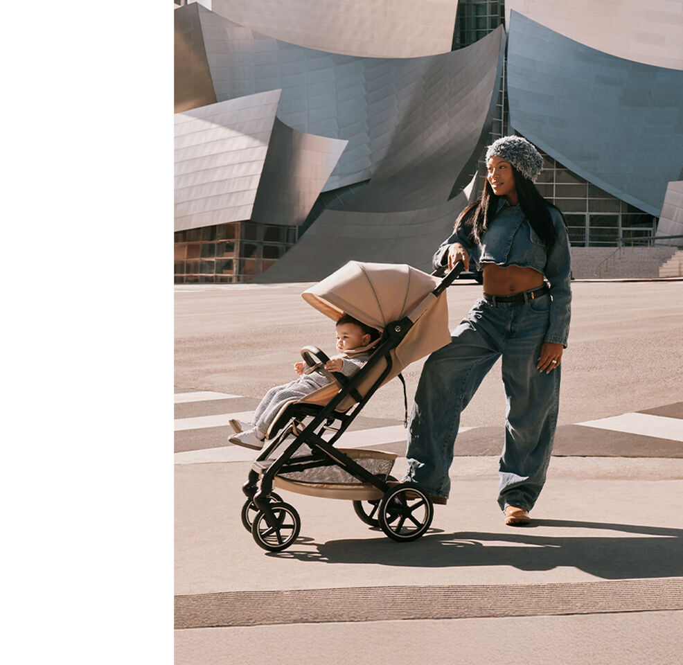 A woman wearing all-denim and a hat with one hand and one foot on her Beezy buggy. Her baby sits inside the buggy.