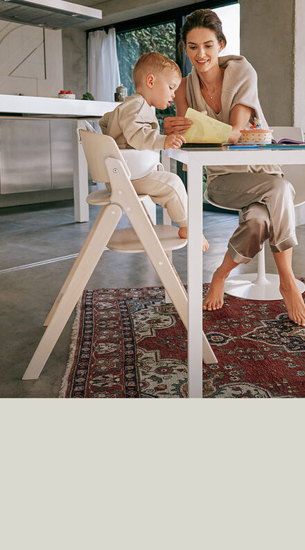 A mother sitting at a table next to her child in a Click & Fold high chair, showing him something on a piece of paper.