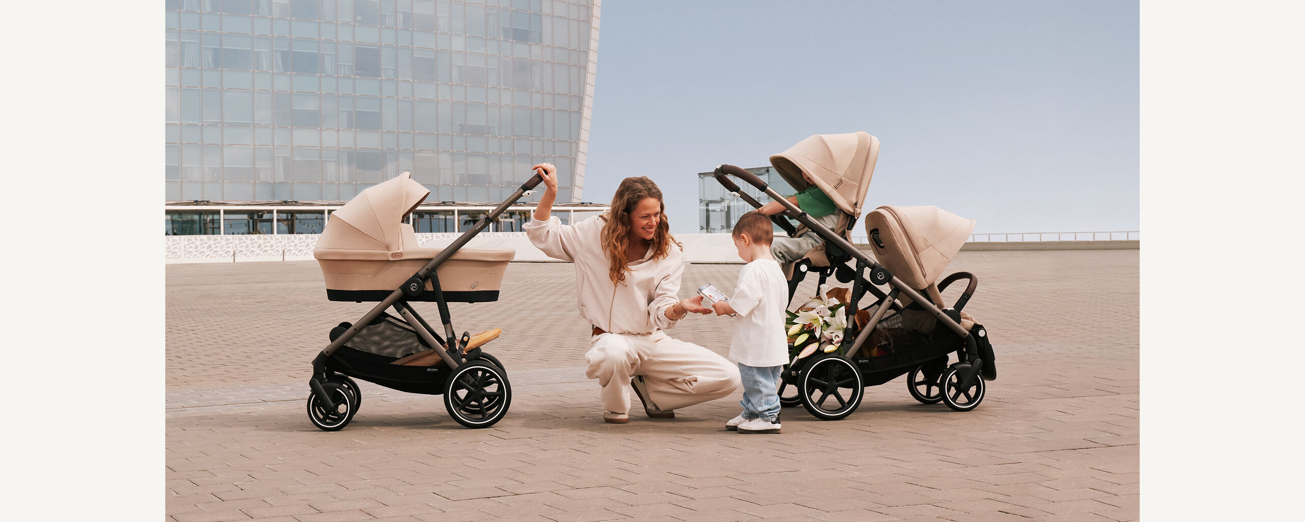 A mother is kneeling on the ground next to her child, grinning at him. She has two Gazelle S strollers next to her, one in single mode, one in sibling mode.