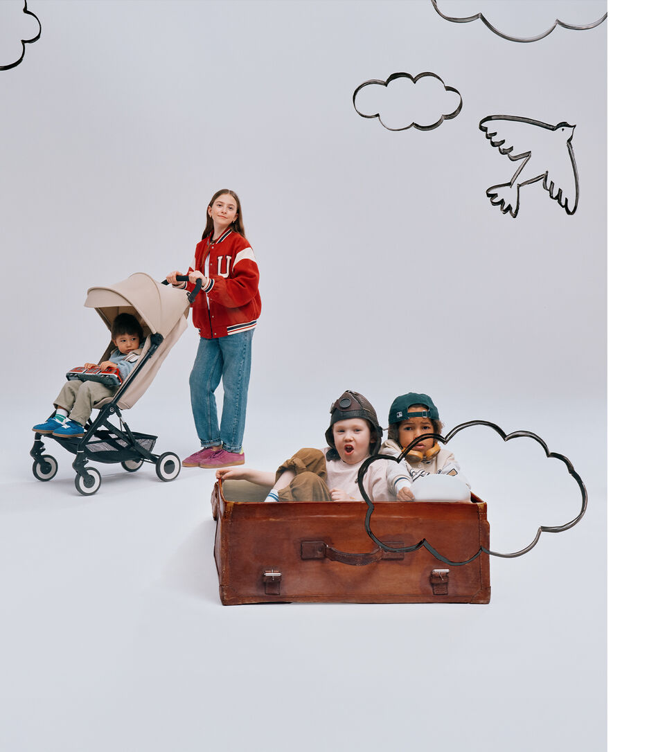 A young girl smiling into the camera as she pushes another child in the Agis buggy. In front of her, there are two more children playing inside a bag as if they were on an airplane.