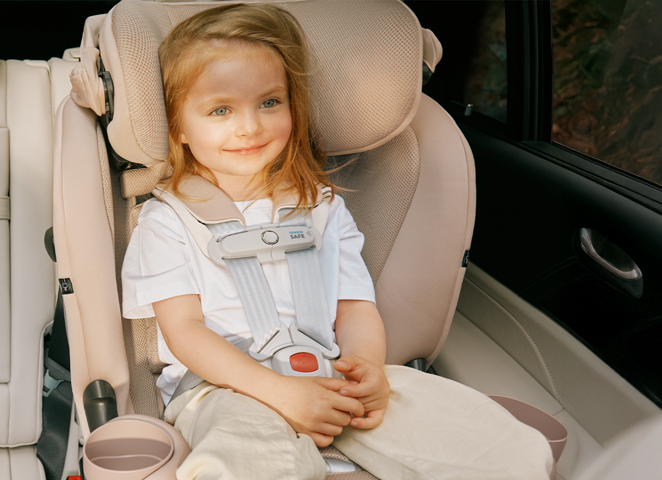 A child happily sits in a CYBEX car seat with a SensorSafe clip attached to the harness.