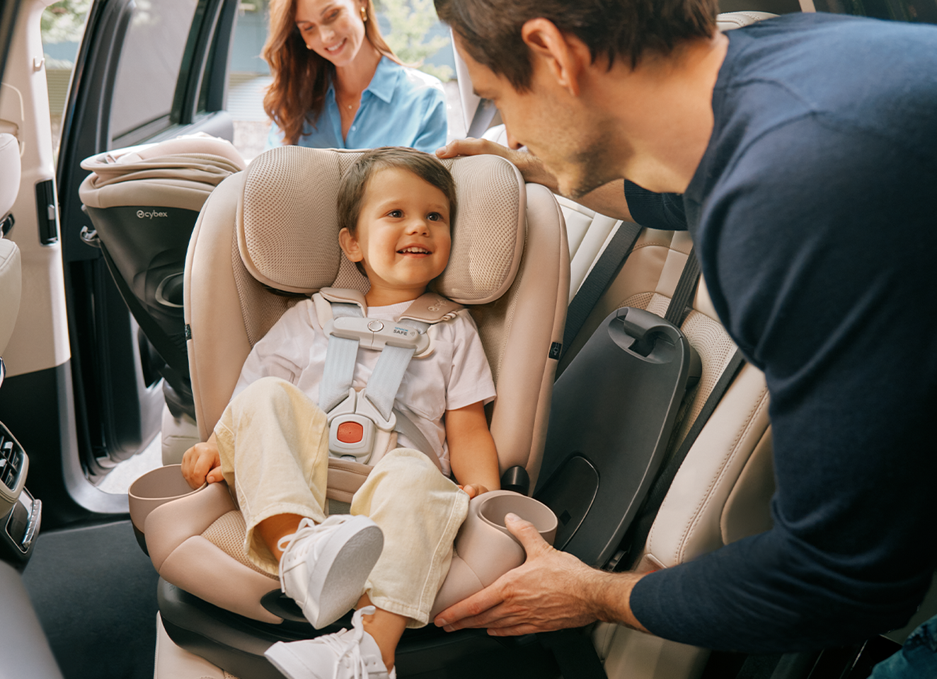 A smiling toddler sits in a car seat, the father in front of him, the mother behind with another CYBEX car seat.