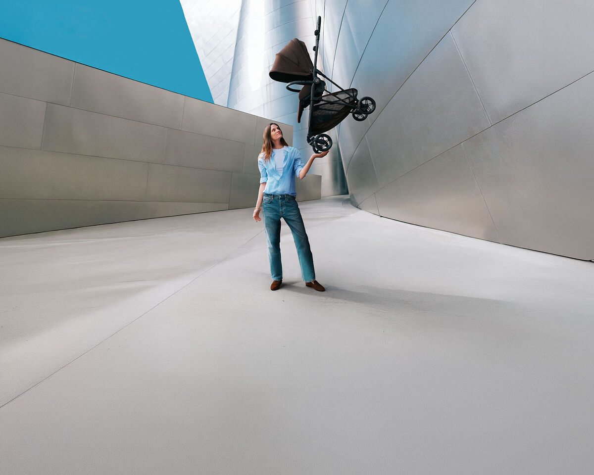 A mother in between two Gazelle S strollers. On the left is one with a cot attached, on the right one with two seat packs. She is holding out her hand to her son who is standing in front of her and handing her something.