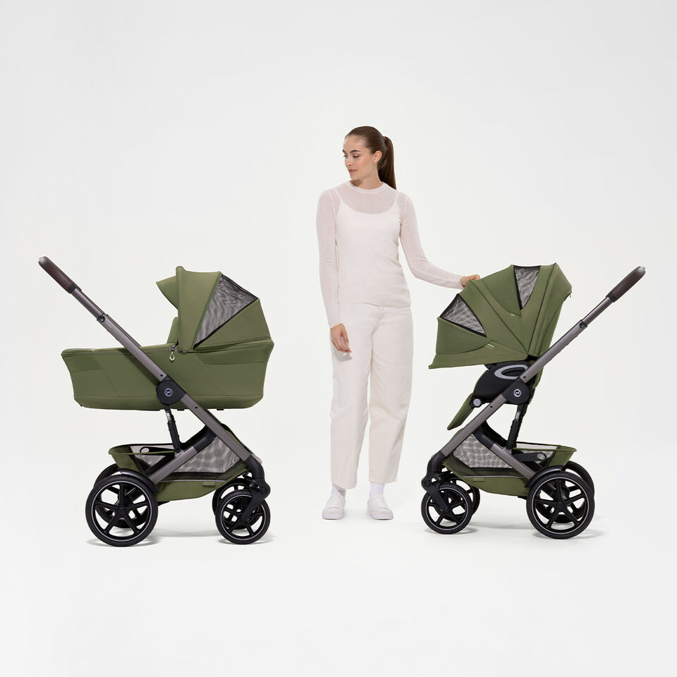 A woman standing between two Talos S Lux strollers. On the left, one with the Cot, on the right, one with the seat pack.