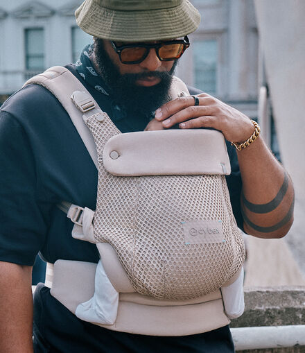 A man wearing a hat and sunglasses, one hand on the top of his child's head that is sitting in an Amya baby carrier.