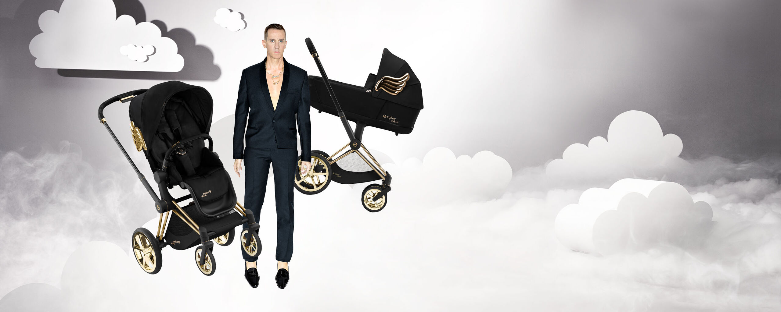 A man in a dark suit stands next to two pram models by Jeremy Scott Wings, surrounded by clouds.
