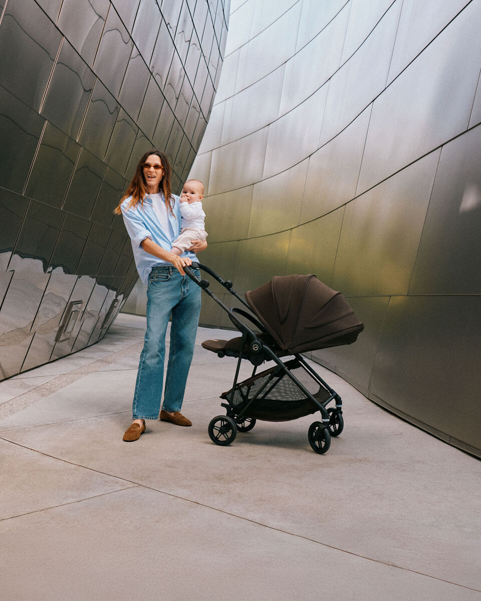 A mother stands with her toddler in her arms and one hand on the handlebar of her Melio Carbon stroller in a narrow passage. The Melio Carbon has its sun canopy down and is in a lie-flat position.