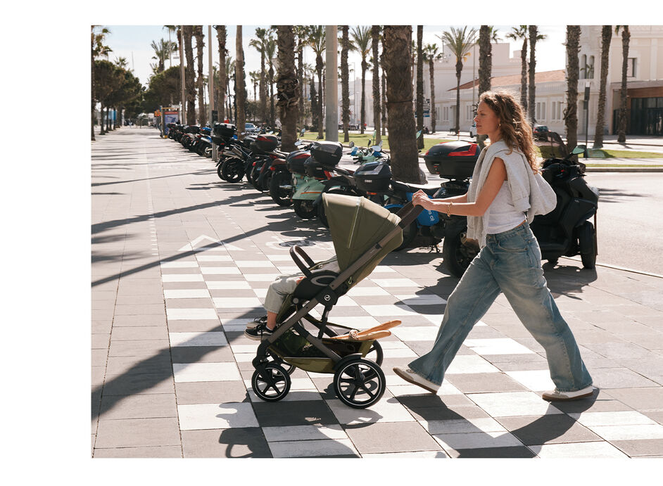 A mother pushing her child in a Talos S Lux stroller around a city. Behind her, multiple motorcycles. 