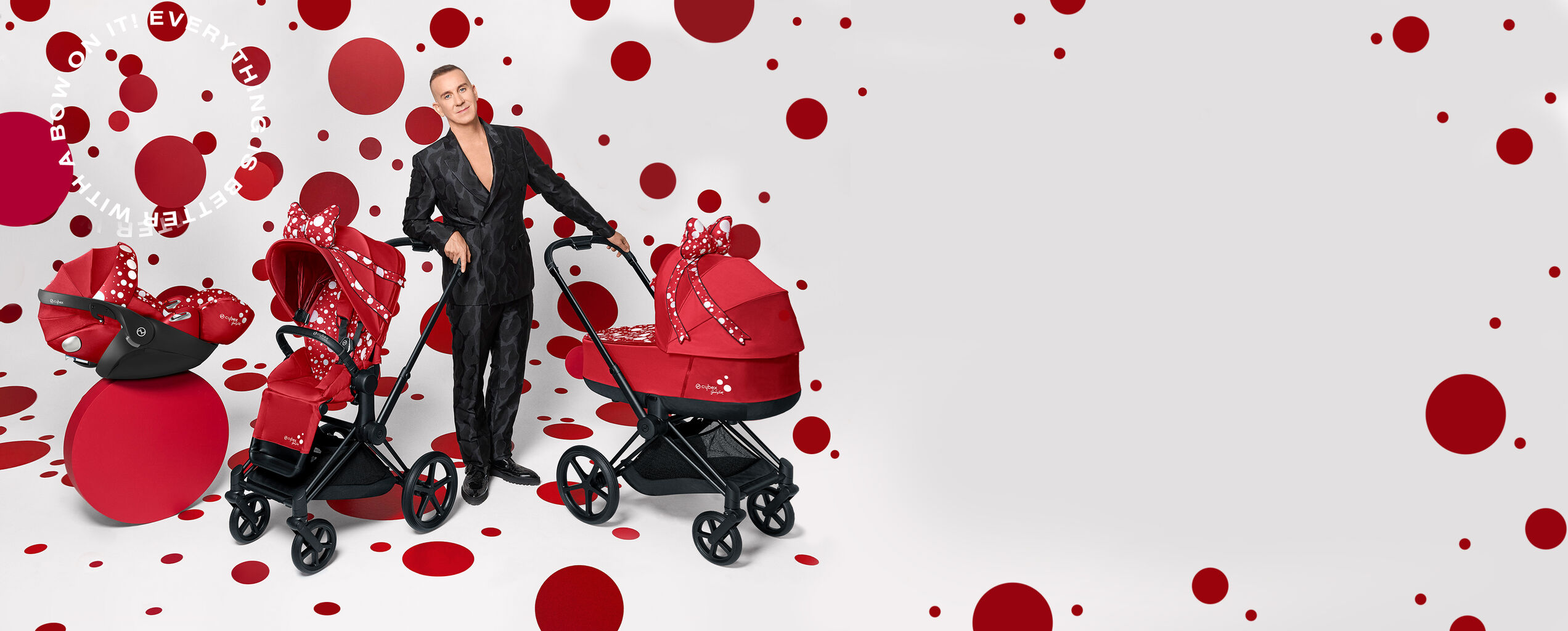 A man in a sleek black suit stands next to three red prams with white polka dots on a background with red dots.