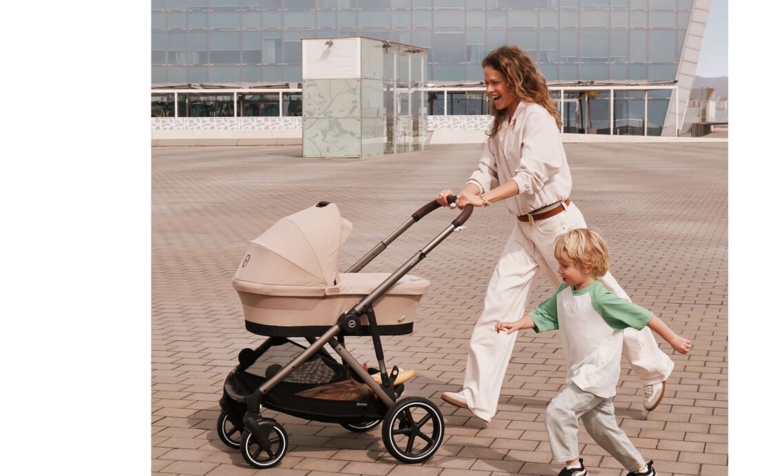 A woman pushing her Gazelle S stroller with a Cot, laughing as she is running together with the son next to her.