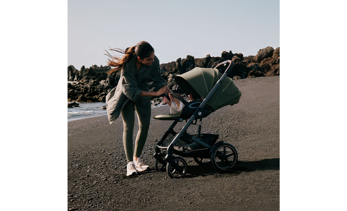 A mother checks on her baby in a Talos S Lux with the seat reclined. They're standing on a stony beach next to the ocean.