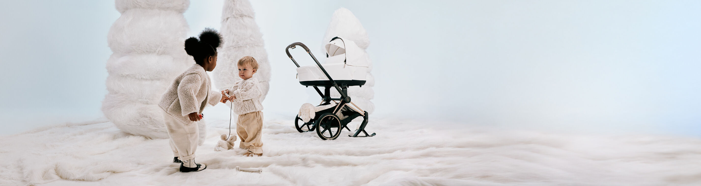 A little girl and boy playing with a toy. Behind them are makeshift white Christmas trees and a Priam with skis attached.