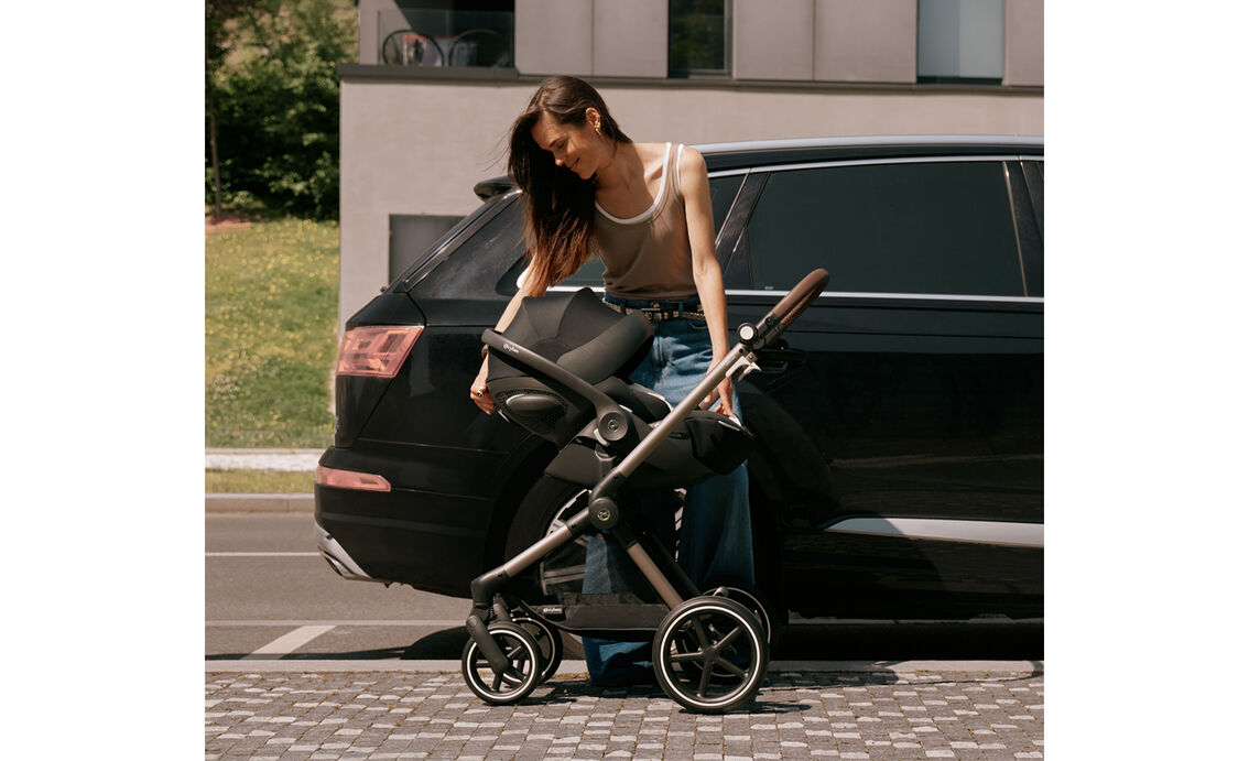 A brunette woman stands beside a black SUV, leaning over a CYBEX stroller frame with an attached infant car seat. The sun canopy of the seat is folded down.