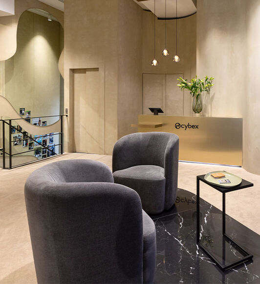 CYBEX store interior with two grey anthracite armchairs facing each other, a sleek slim coffee table, bouquet of flowers and beige walls. Three stylish lamps provide atmospheric lighting.