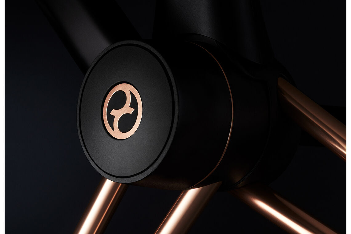 A close-up shot of a dark-coloured round part with a golden logo resembling a stylized letter on it, against a dark and slightly out-of-focus background.