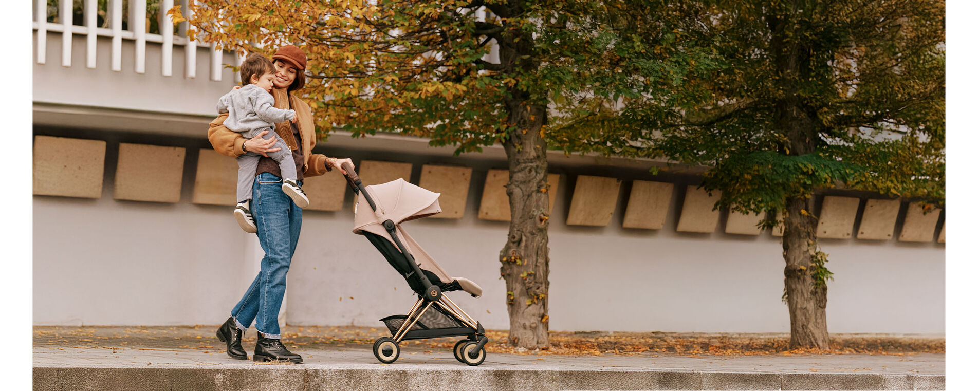 A woman pushing her CYBEX Coya Buggy along in autumn, holding her child in her right arm and smiling at him.