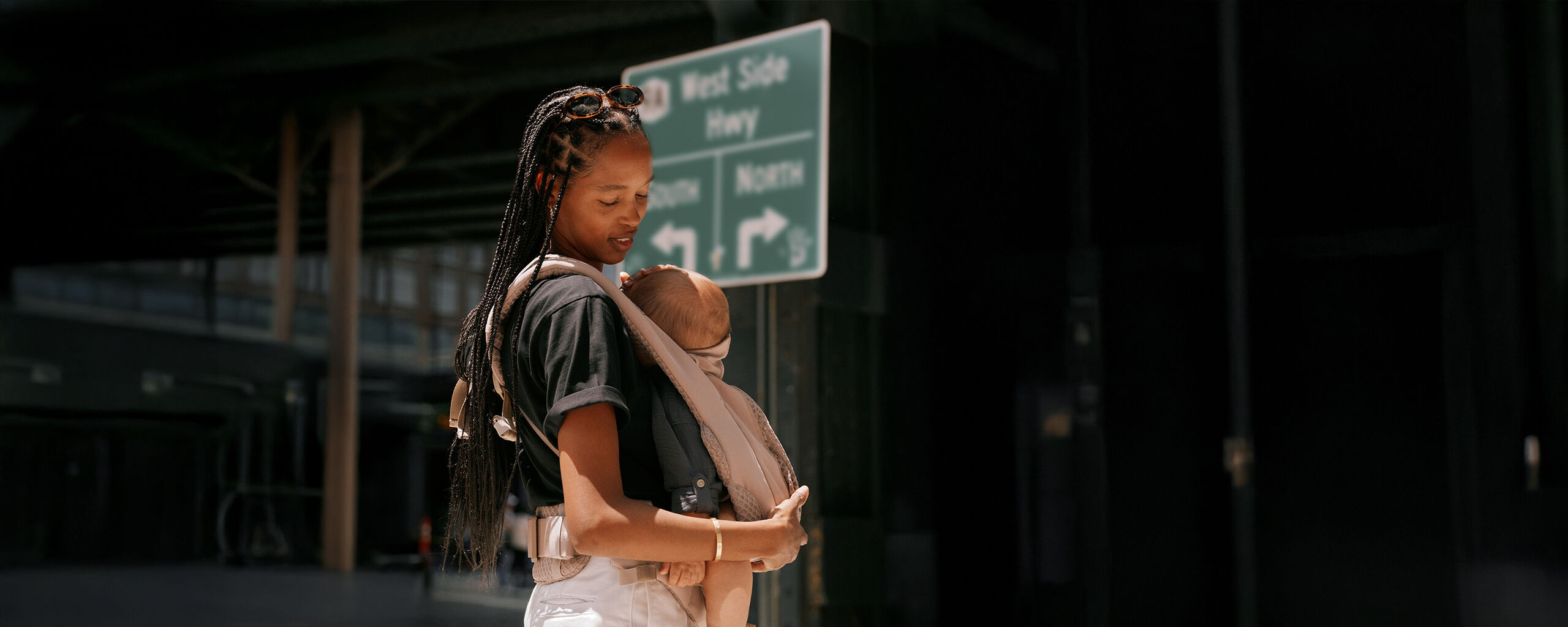Žena s pletenicemi nos&iacute; d&iacute;tě v nos&iacute;tku Coya vedle dopravn&iacute; značky.