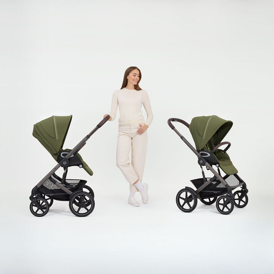 A woman standing next to two Talos S Lux strollers, one with a forward facing seat unit, one with a rear facing one.