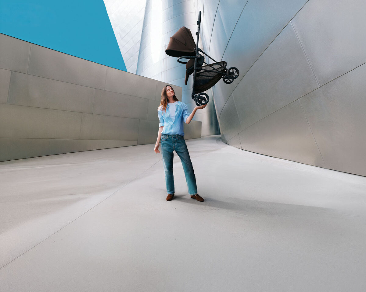 Mujer entre dos cochecitos Gazelle S. A la izquierda hay uno con un capazo acoplado y a la derecha otro con dos seat packs. Ella le tiende la mano a su hijo, que est&aacute; de pie delante de ella y le entrega algo.