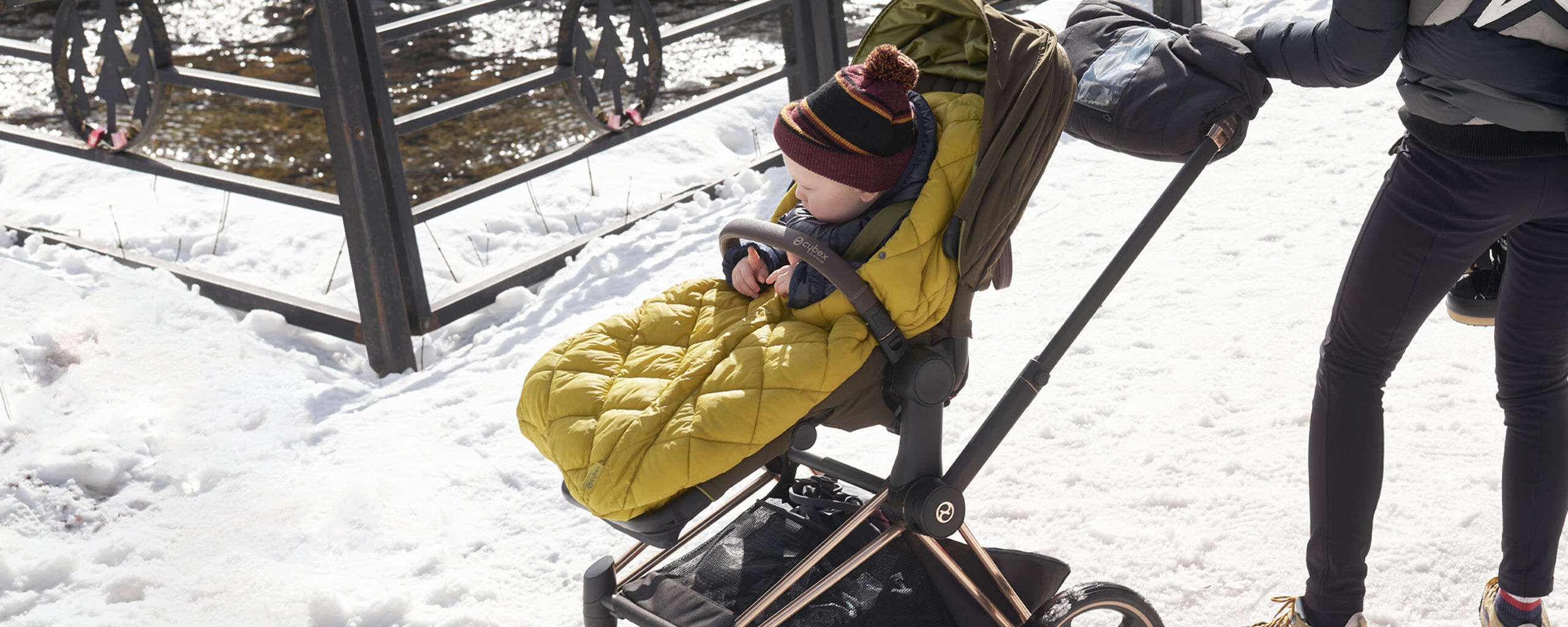 A toddler sits in a yellow footmuff in a pram on a snowy path.