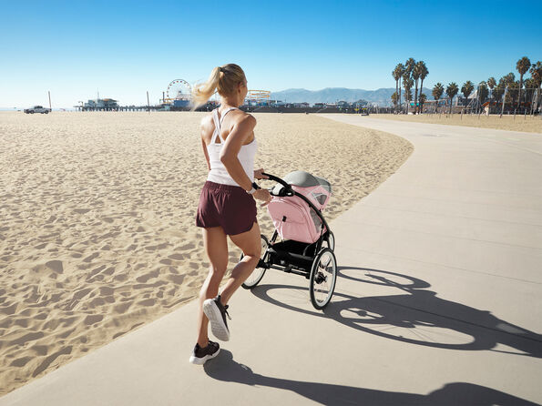 A blonde ponytail woman in white athletic wear and burgundy shorts runs while pushing a pink running pushchair (Zeno) on a cement path parallel to a sandy beach, with palm trees and a Ferris wheel in the background, seen from behind.