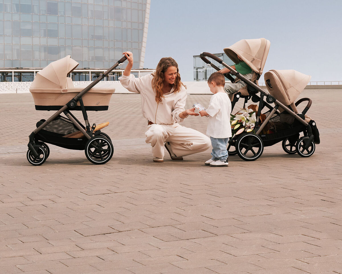 A mother in between two Gazelle S strollers. On the left is one with a cot attached, on the right one with two seat packs. She is holding out her hand to her son who is standing in front of her and handing her something.