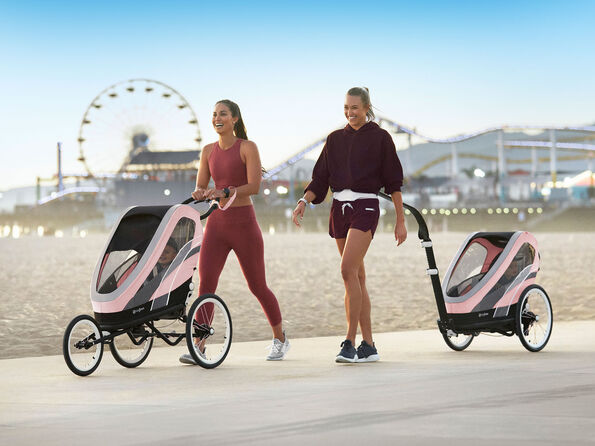Two women pushing Zeno jogging pushchairs along a beach, with a Ferris wheel in the background.