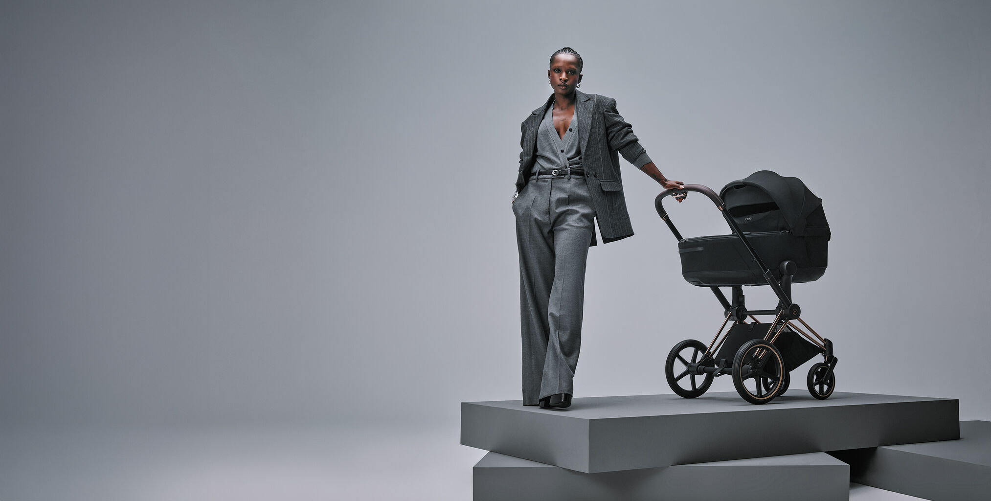 A woman wearing all gray with one hand in her pants pocket and the other holding the Priam handlebar casually, looking directly into the camera.