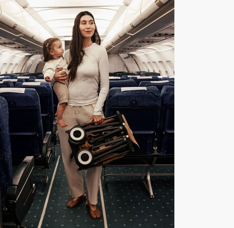 A mother on a plane amidst plane seats, holding her daughter with one hand and a folded Libelle with the other.