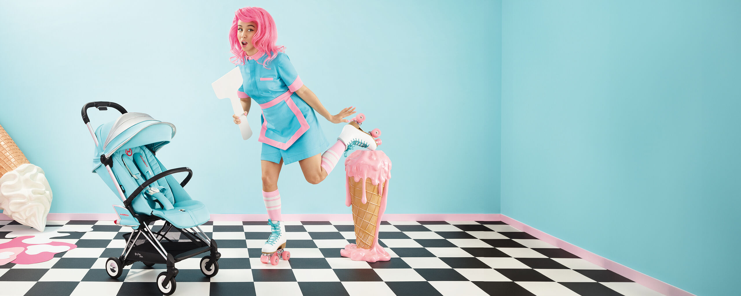 A woman with a pink wig and roller skates poses in a pastel blue room next to a stroller and a large ice cream cone.