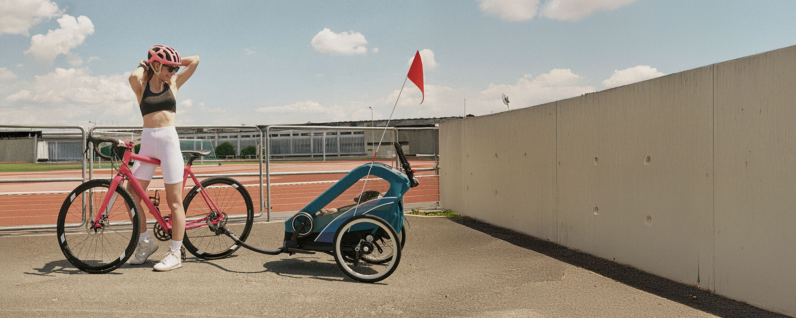 Une femme pr&egrave;s d&rsquo;une piste de running, attachant sa queue de cheval et se retournant pour voir son enfant assis dans sa Zeno Bike.